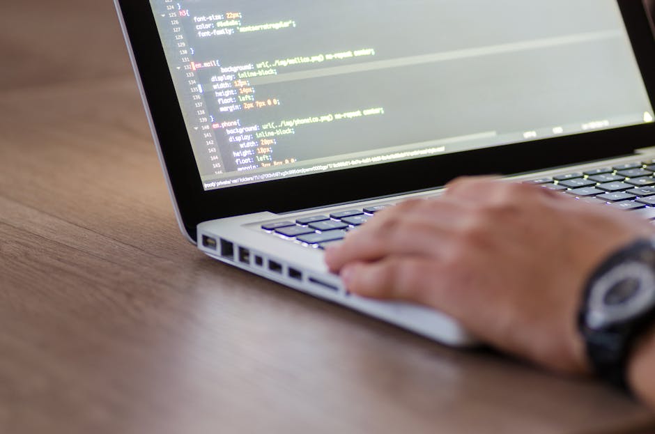 A close-up view of a person using a silver laptop placed on a wooden desk, with the right hand poised over the keyboard. The laptop screen displays lines of colorful code or script, suggesting program